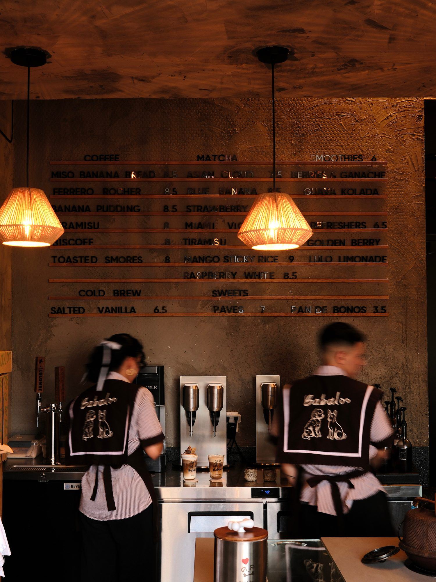 Two Babaloo baristas in striped aprons working behind the counter under the menu board